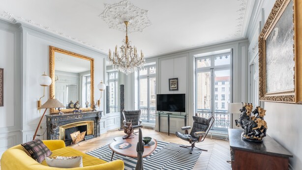 A spacious living room with a yellow sofa, a fireplace, a chandelier, and a large window with a view of buildings. - Célestins - Haut standing 120m² - Coeur de Lyon (Lyon)
