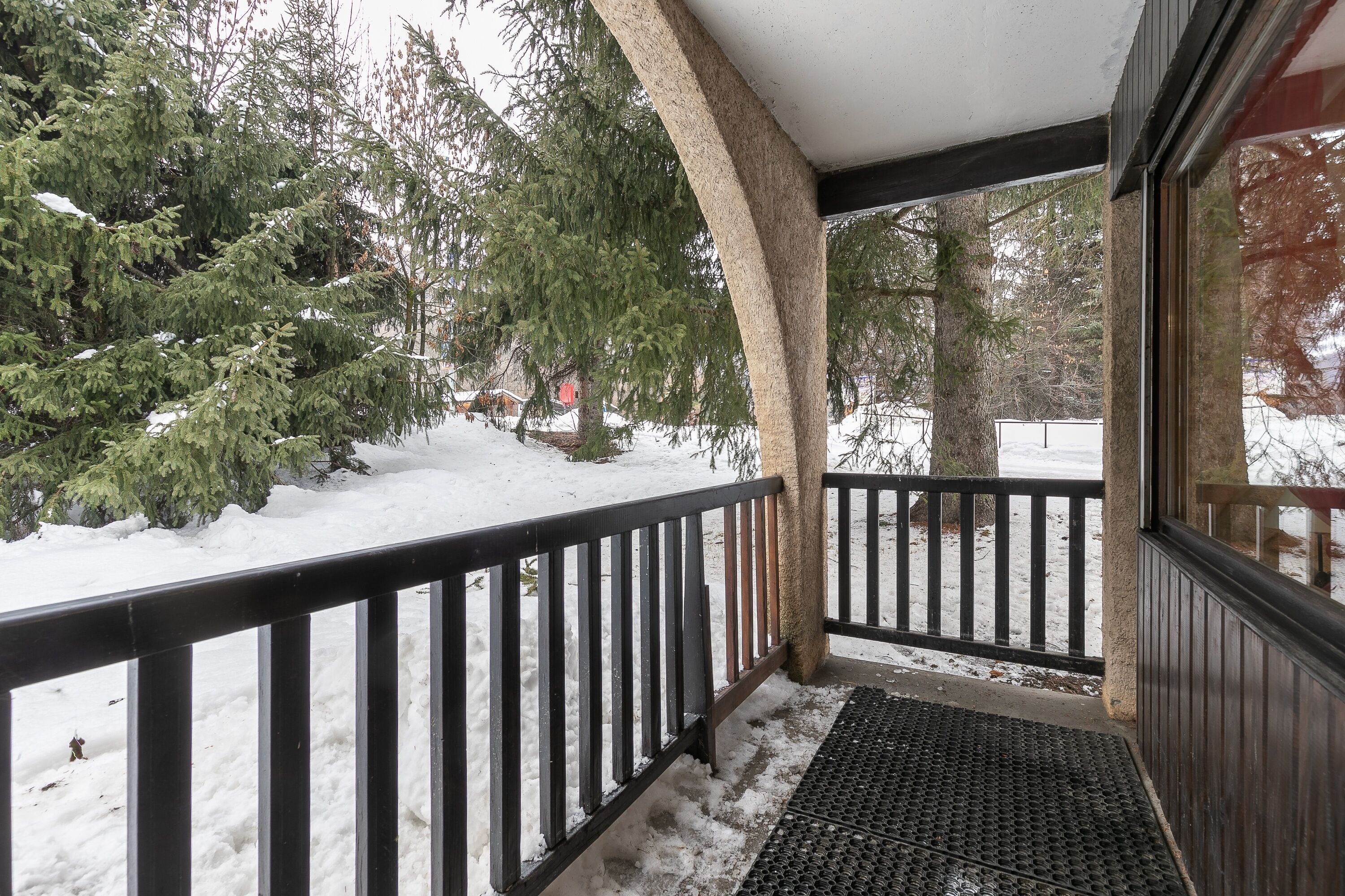 A snow-covered porch with a black railing, a wooden bench, and a view of snow-laden trees and a house in the background.