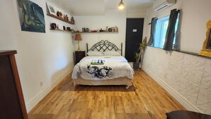 Une chambre à coucher comprenant un lit, un plancher en bois, un luminaire au plafond, une étagère fixée au mur avec des objets décoratifs, et une porte.