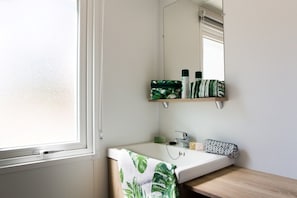 Een badkamer met een witte wastafel, een spiegel en een raam. Er staan toiletartikelen op een plank boven de wastafel.