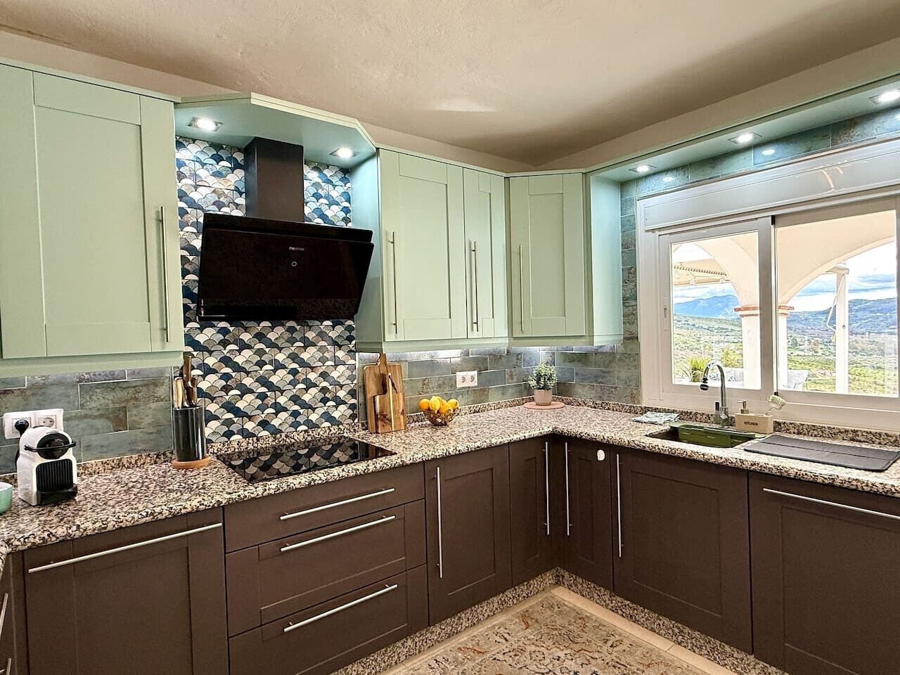 A kitchen with dark cabinets, a granite countertop, a black range hood, and a window with a view.