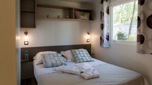A neatly made bed with pillows and a folded towel, a bedside table with a water bottle, and a window with curtains. - Bréhadour - Confort 4 TV - 9p (7) (Guérande)