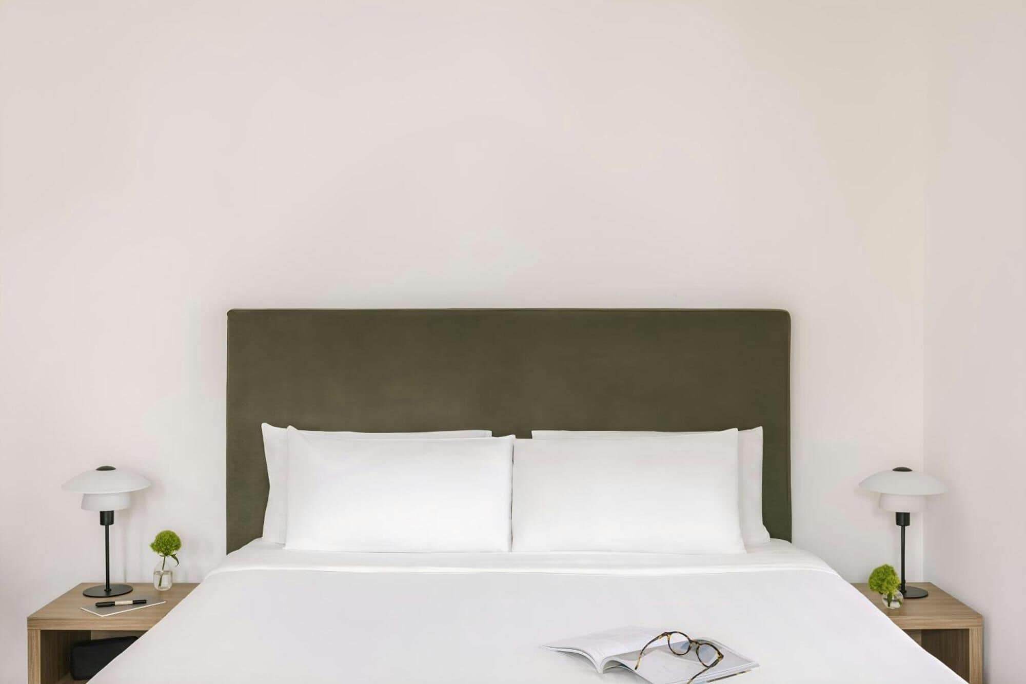 A neatly made bed with white linens and a green headboard, flanked by two bedside tables with lamps and small plants.