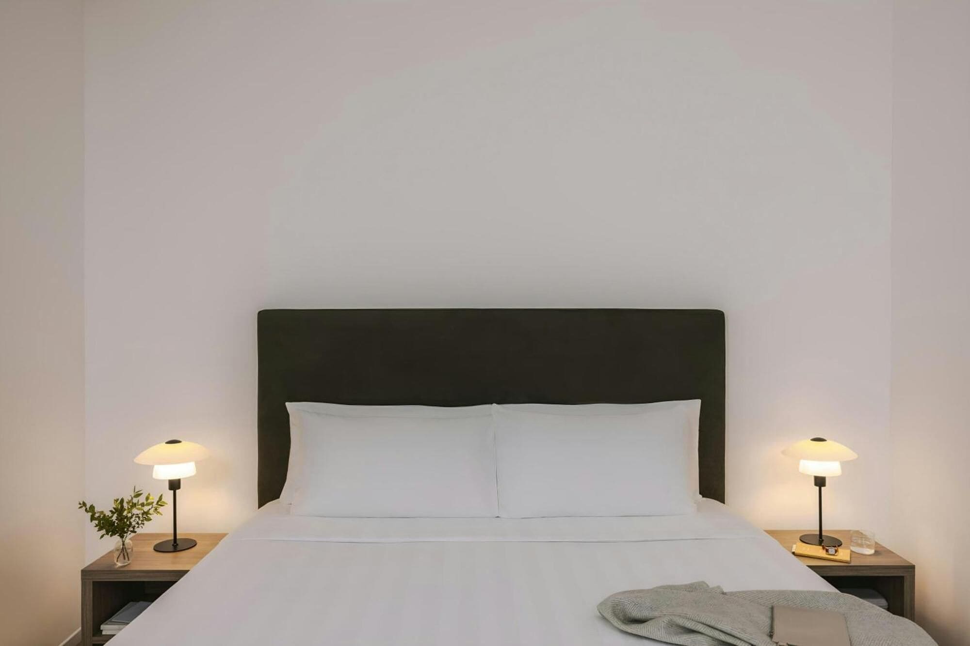 Una cama bien hecha con sábanas blancas, un cabecero oscuro y dos mesitas de noche con lámparas.
