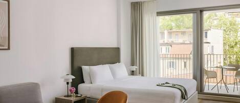 A hotel room with a bed, a desk, a chair, a lamp, and a view of the outside.