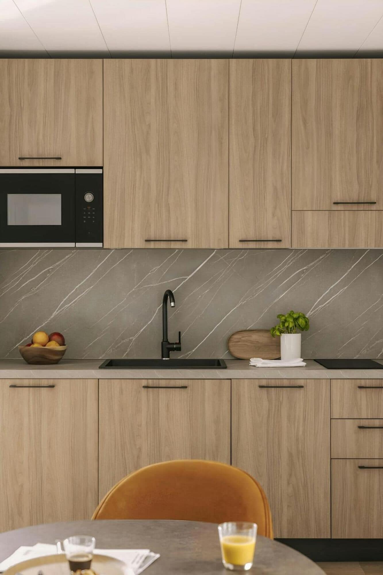 Une cuisine moderne avec des meubles en bois, un four à micro-ondes encastré, un pare-feu en marbre, un évier avec un robinet noir et un coin repas avec une table et des chaises.