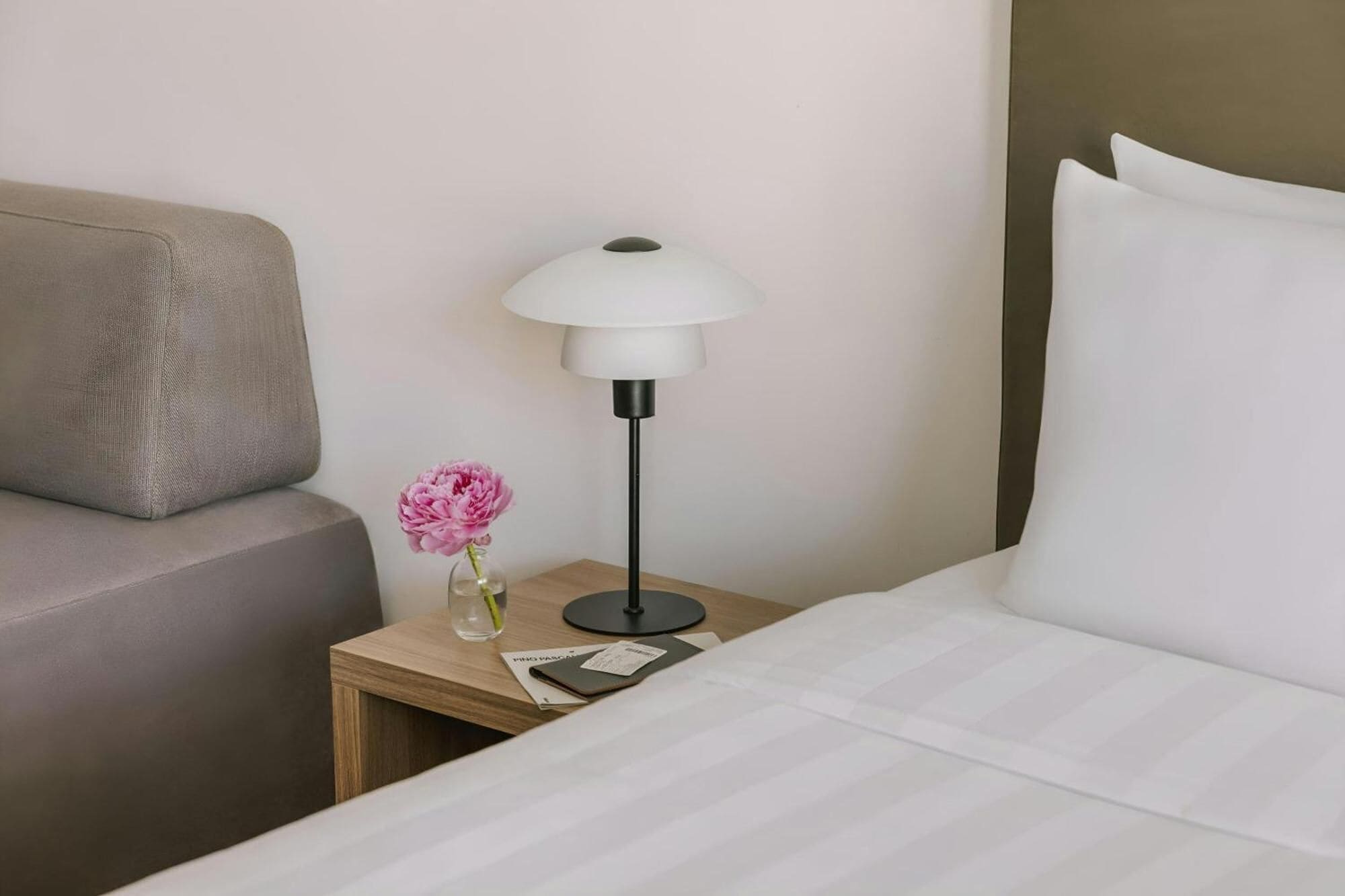 A hotel room with a bed, a nightstand, a lamp, a vase with a flower, and a sofa.