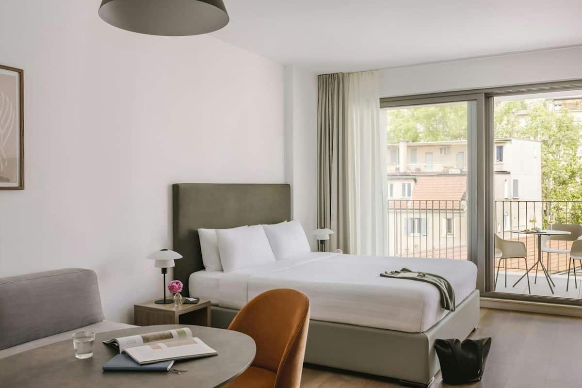 Habitación de hotel con cama, escritorio, silla, lámpara y vistas al exterior.