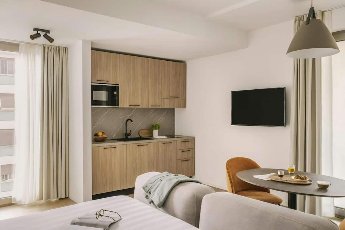 Una habitación de hotel moderna con kitchenette, televisor de pantalla plana, zona de comedor con mesa y sillas, y cama con vista a la ciudad.