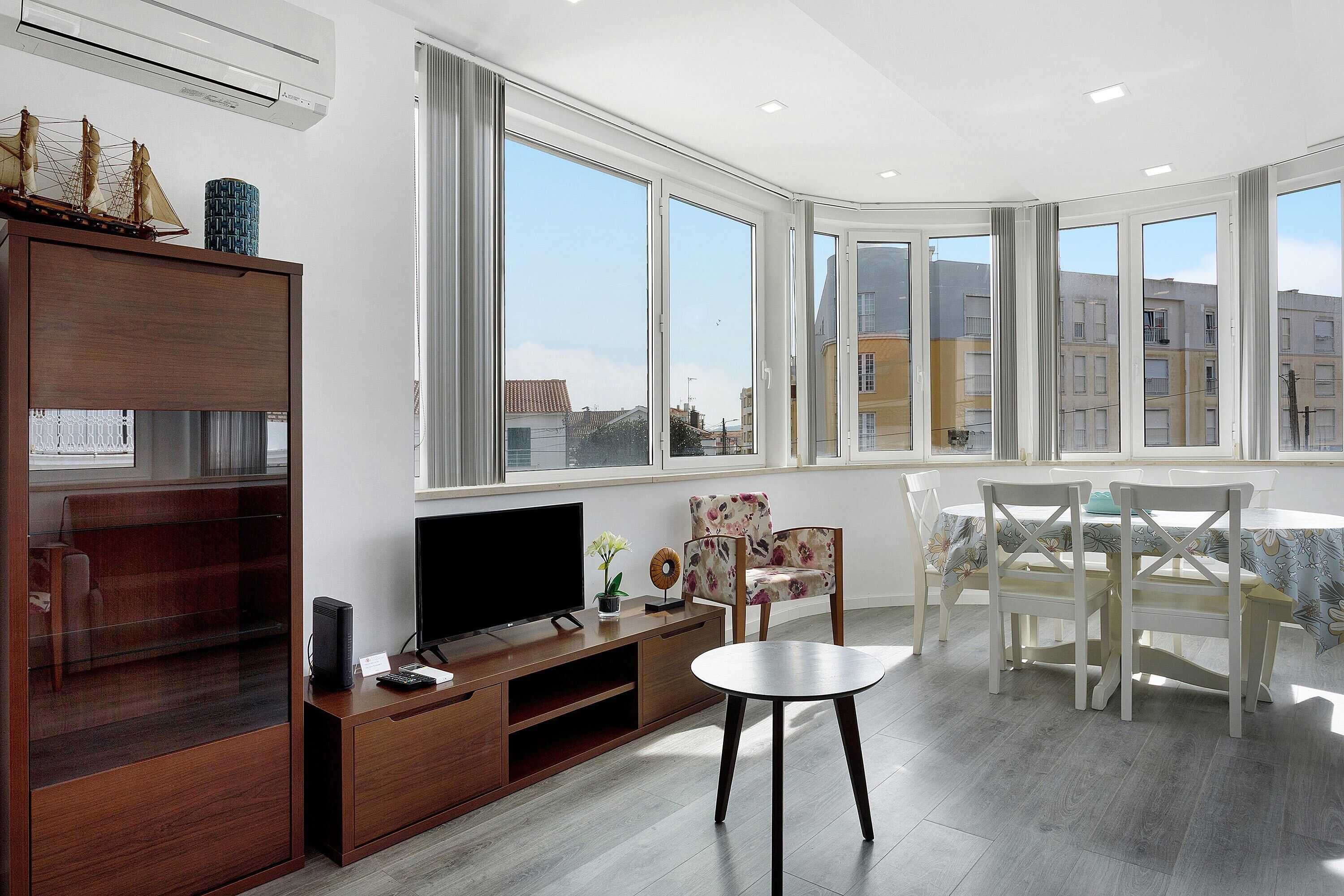 A modern living room with a TV, wooden entertainment unit, dining table, and large windows offering a city view.