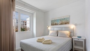 Un dormitorio con cama, mesita de noche, ventana y un cuadro en la pared.