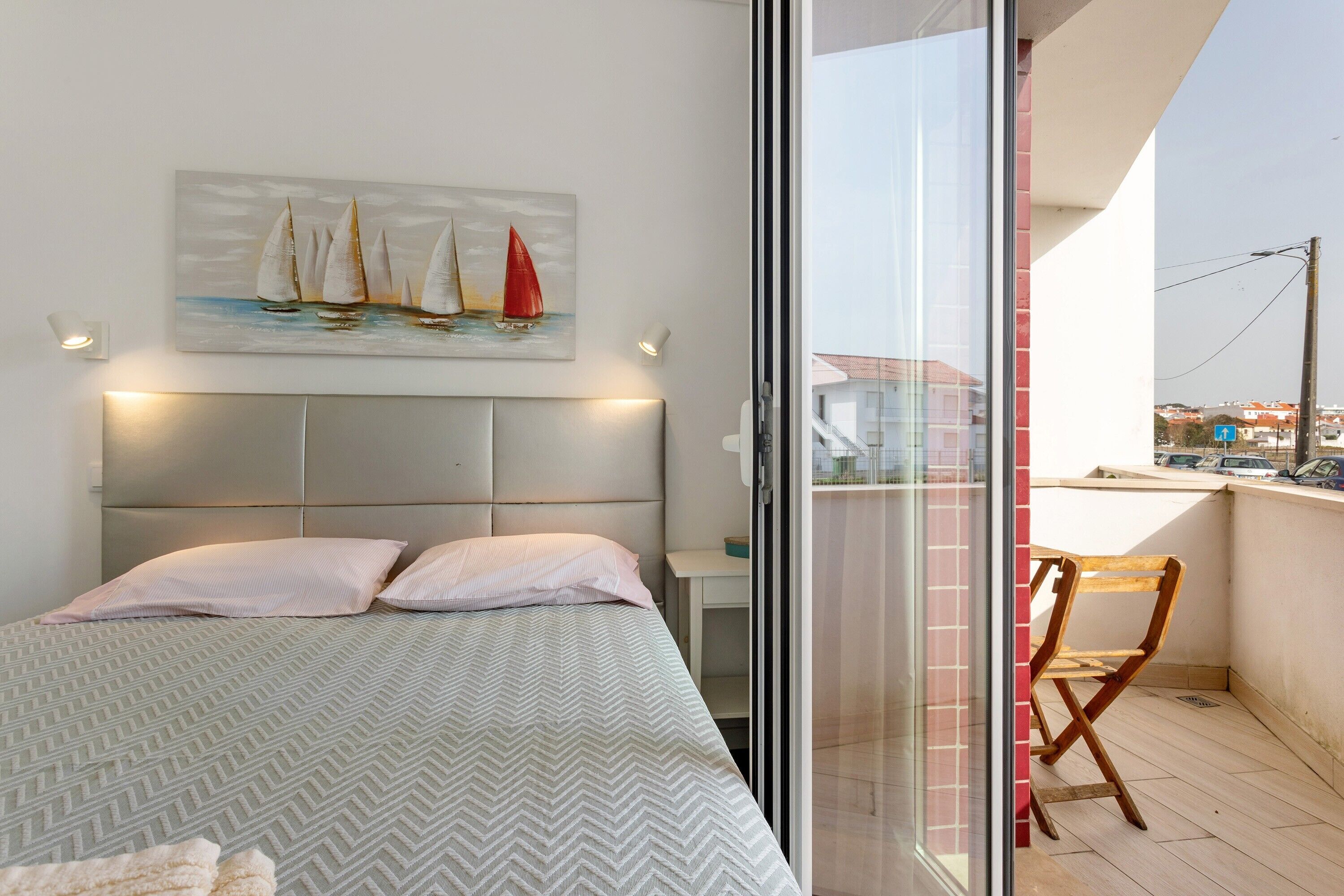 A bedroom with a bed, pillows, a nightstand, a painting of sailboats, and a view of the city through a glass door.