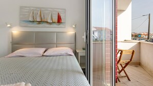 Un dormitorio con cama, almohadas, una mesita de noche, un cuadro de veleros y vista de la ciudad a través de un ventanal.