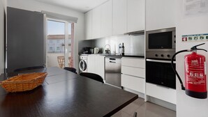 Una cocina moderna con armarios blancos, electrodomésticos de acero inoxidable, una mesa de comedor con una cesta y un extintor de incendios en la pared.