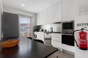 A modern kitchen with white cabinets, stainless steel appliances, a dining table with a basket, and a fire extinguisher mounted on the wall. - Apartment 'Casa Dos Terraços T2 Rc' with Balcony, Wi-Fi and Air Conditioning (São Martinho do Porto)