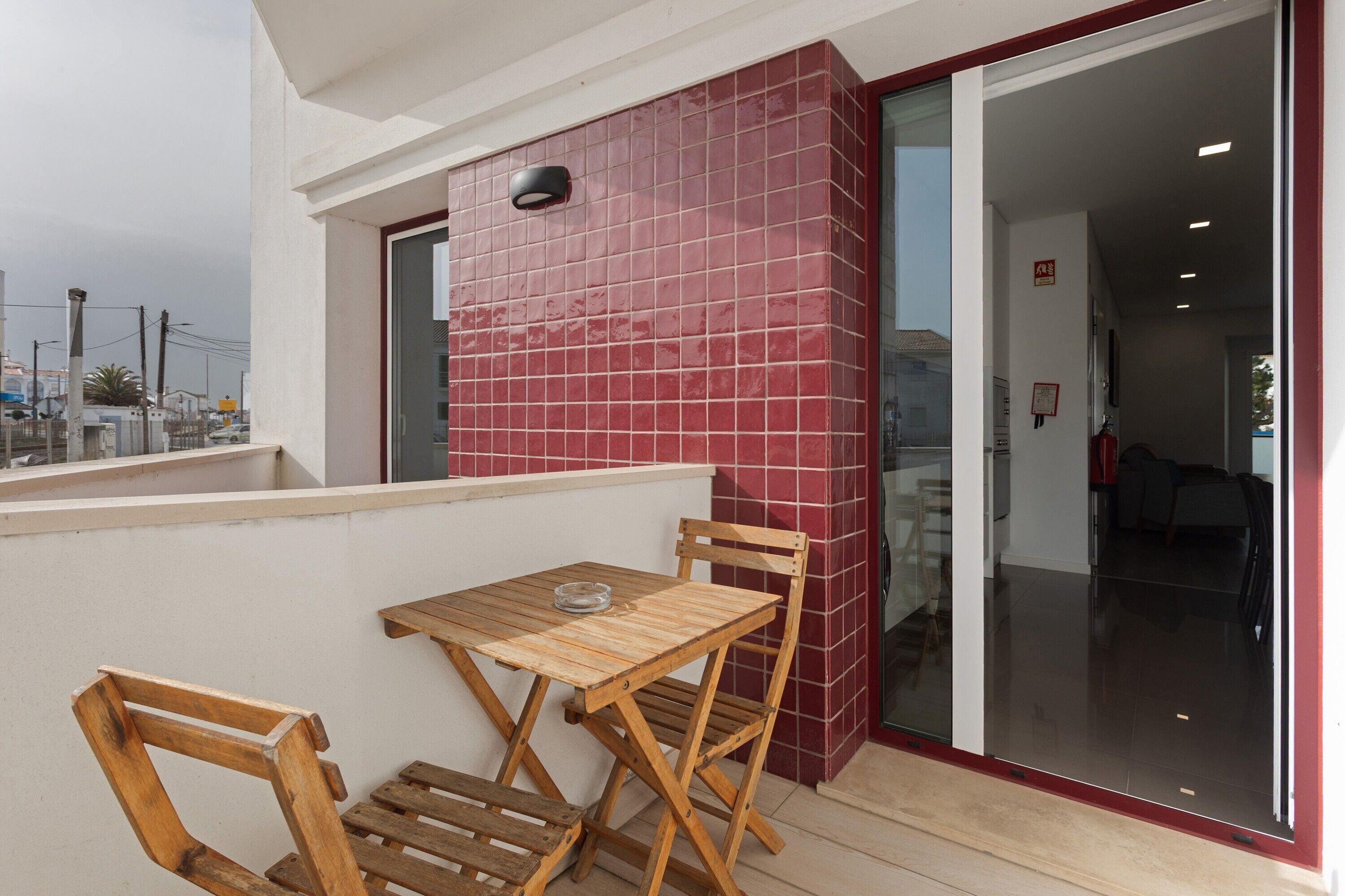 A balcony with a wooden table and chairs, a red tiled wall, and a glass door leading inside.