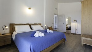 Een net opgemaakt bed met een blauwe spreideken, witte kussens en opgerolde handdoeken. Er staat een bedlampje, een houten nachtkastje en een houten garderobe.