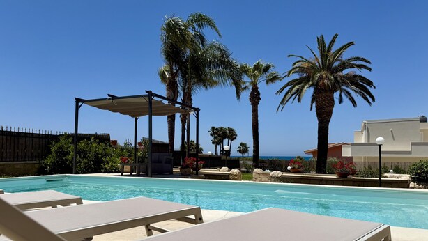 Een zwembad met ligbedden, een pergola, palmbomen en uitzicht op zee.