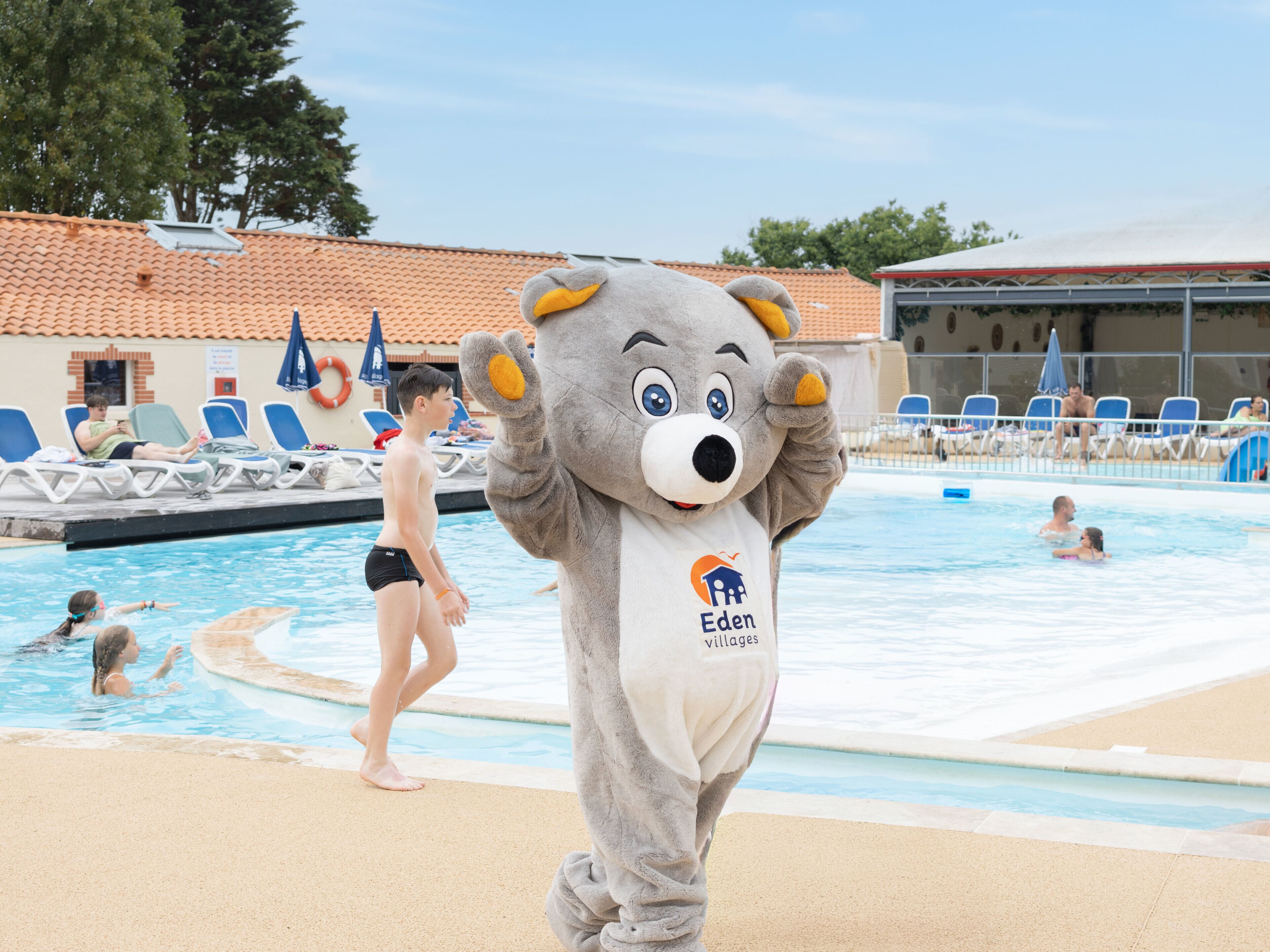 Un personaje promocional cerca de una piscina con sillas de descanso y un techo de azulejos al fondo.
