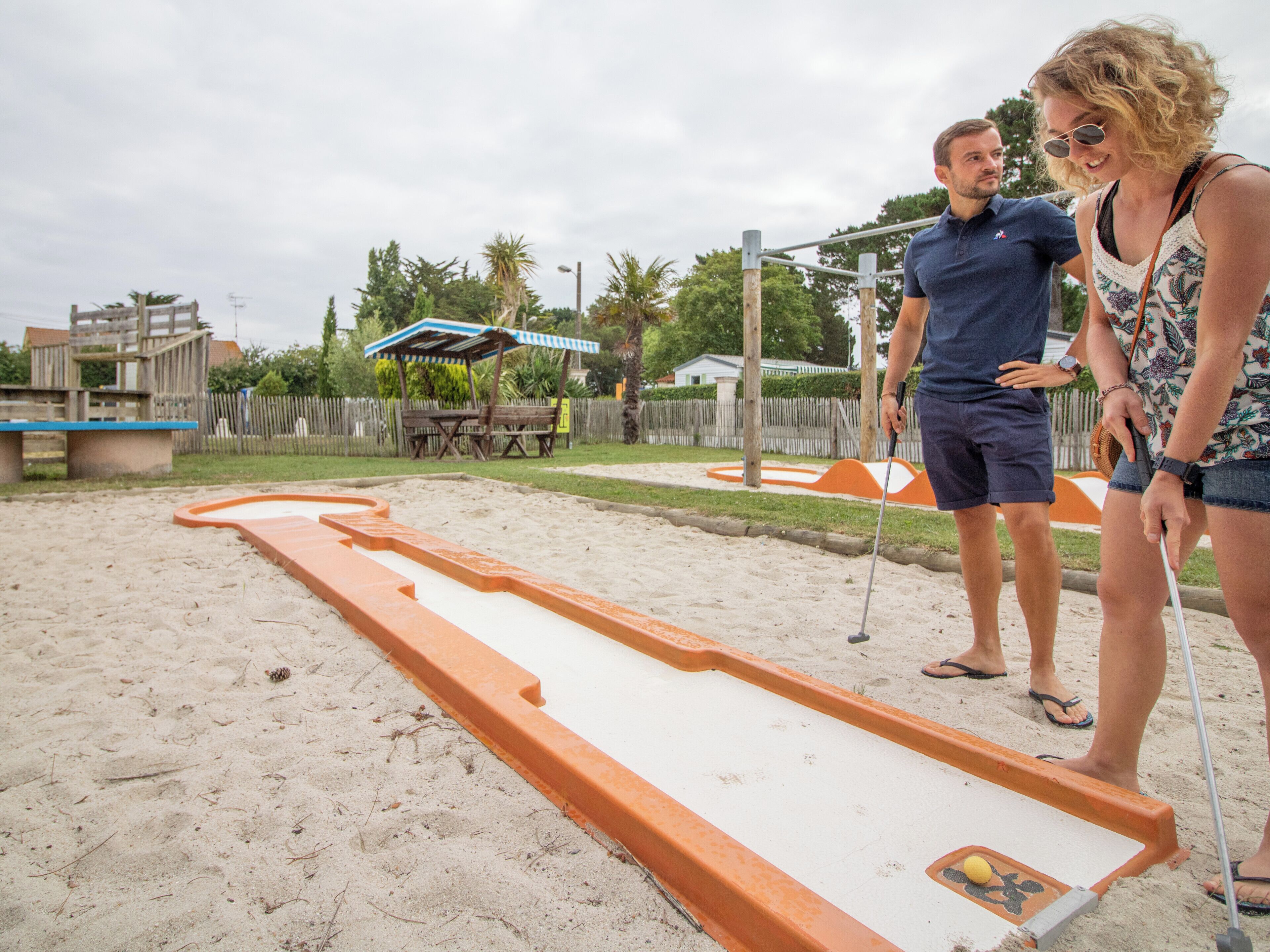 Un terrain de sable avec une piste longue et étroite aux couleurs orange et blanche, un banc en bois et, au fond, un auvent rayé de bleu et de blanc.