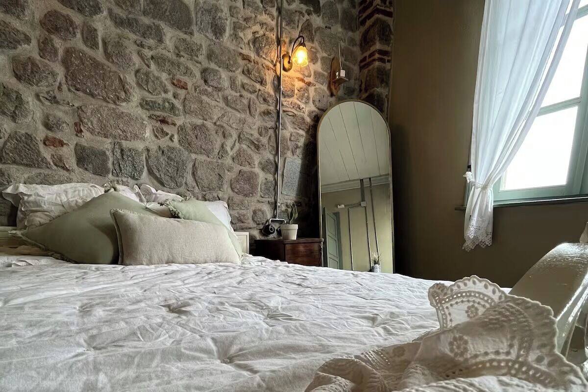 A bedroom with a stone wall, a bed with white bedding, a nightstand with a lamp, and a window with sheer curtains.