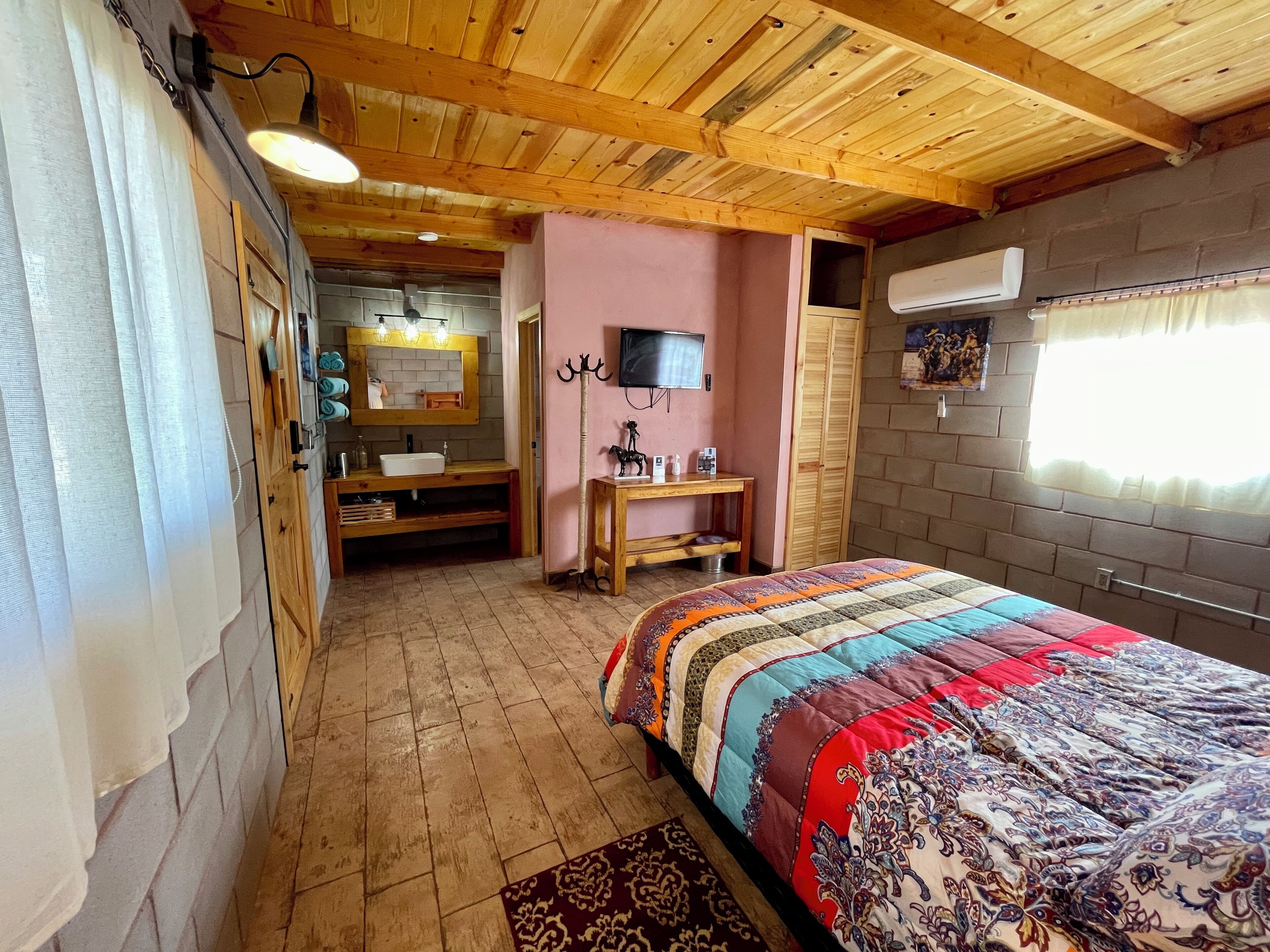 A bedroom with a wooden ceiling, a bed with a colorful quilt, a vanity with a mirror, and a television on the wall.