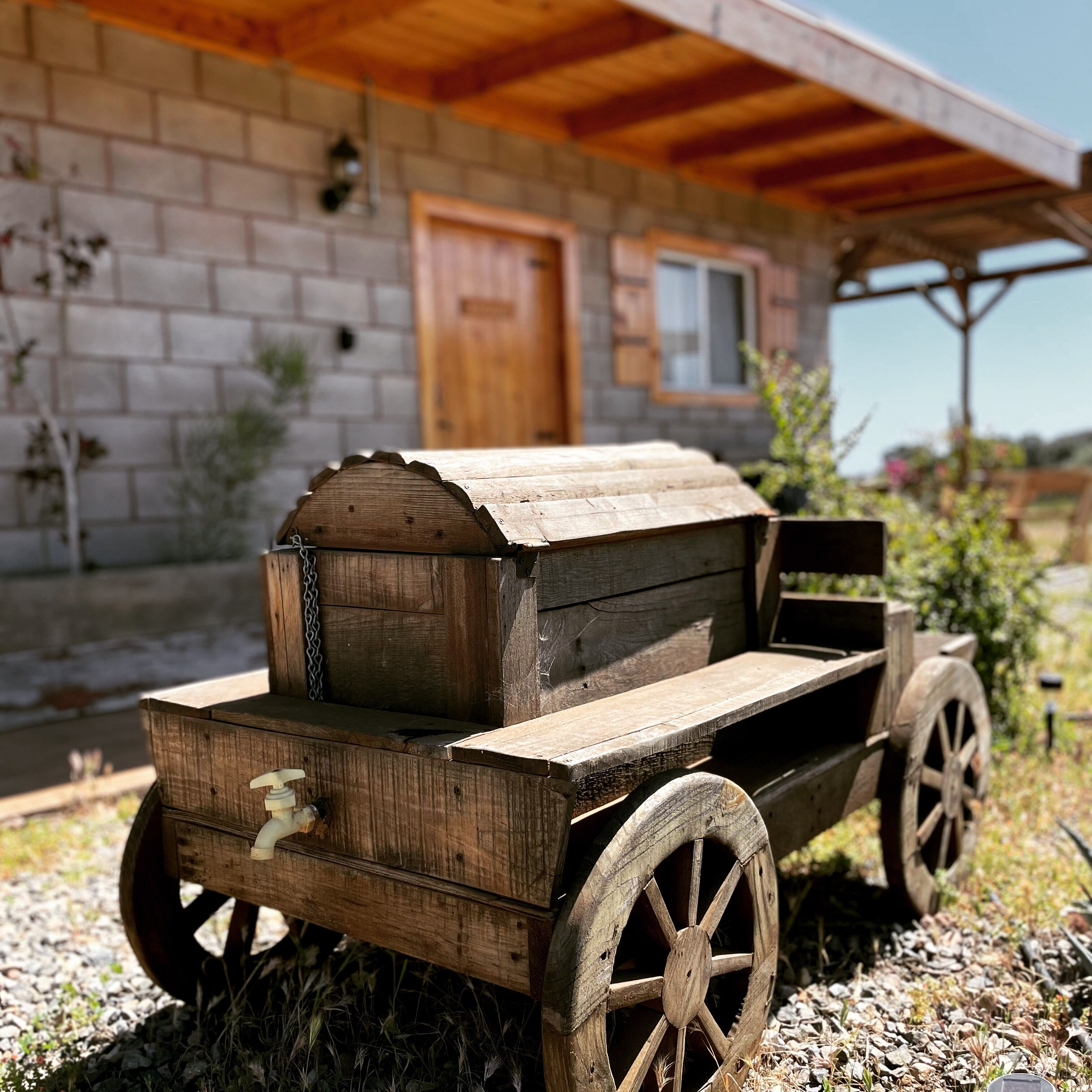 Une charrette en bois rustique, dotée d’une plateforme plate, est placée devant un bâtiment en pierre avec une porte et une fenêtre en bois.