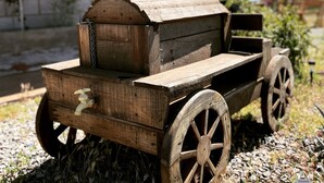 Un carretto di legno rustico con rimorchio piatto, posizionato di fronte a un edificio in pietra con porta e finestra in legno.