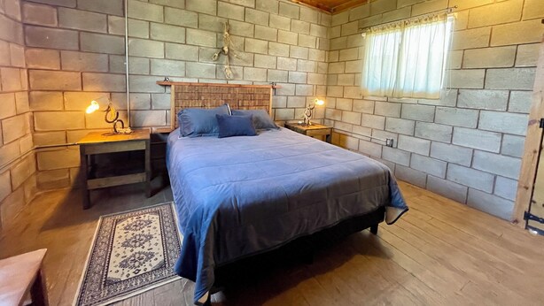 A bedroom with a wooden ceiling, brick walls, a bed with a blue bedspread, two bedside tables with lamps, and a window with curtains. - Wine Country Retreat: Sleeps 8-Beautiful 4BR Cabin with A/C  Valle de Guadalupe (Ensenada)