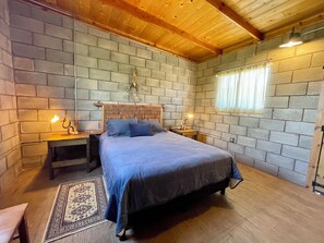 A bedroom with a wooden ceiling, brick walls, a bed with a blue bedspread, two bedside tables with lamps, and a window with curtains. - Wine Country Retreat: Sleeps 8-Beautiful 4BR Cabin with A/C  Valle de Guadalupe (Ensenada)