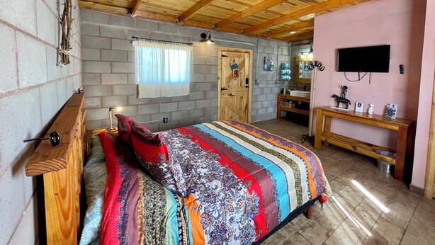 Una camera da letto con soffitto in legno, un letto con coperta colorata, un comò in legno, un comodino con lampada, una finestra con tende e un bagno con lavandino e specchio.