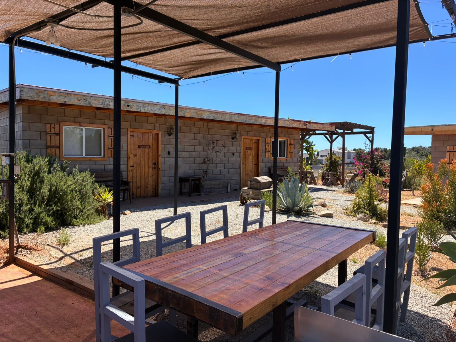 A covered patio with a wooden table and chairs, a stone building with wooden doors, and a garden with various plants.