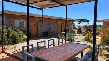 A covered patio with a wooden table and chairs, a stone building with wooden doors, and a garden with various plants.
