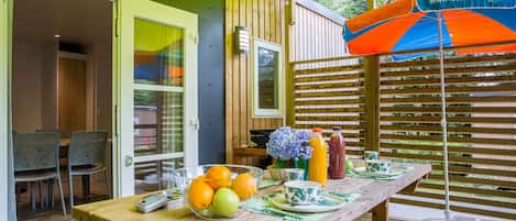 Un espace de restauration en plein air avec une table en bois, dressée avec des fruits, un saladier et un vase de fleurs.