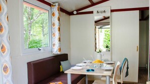 A cozy room with a brown sofa, a dining table with chairs, and a window with a view of trees. - Truffe - Chalet confort 2 – 5p (2) (Saint-Geniès)