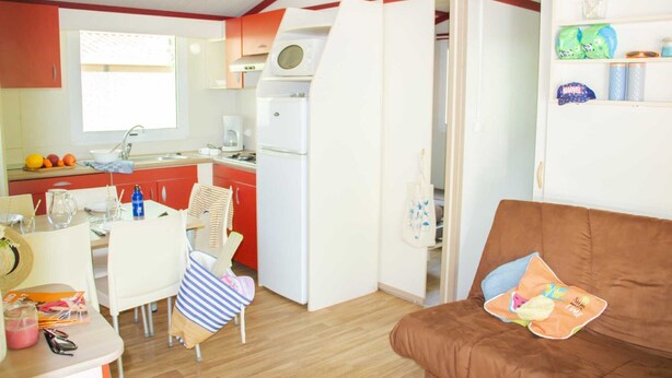 A compact kitchen with a refrigerator, microwave, and sink. A dining table with chairs is positioned near the window. A brown sofa with a cushion is in the foreground. - Truffe - Chalet confort 3 – 6p (1) (Saint-Geniès)