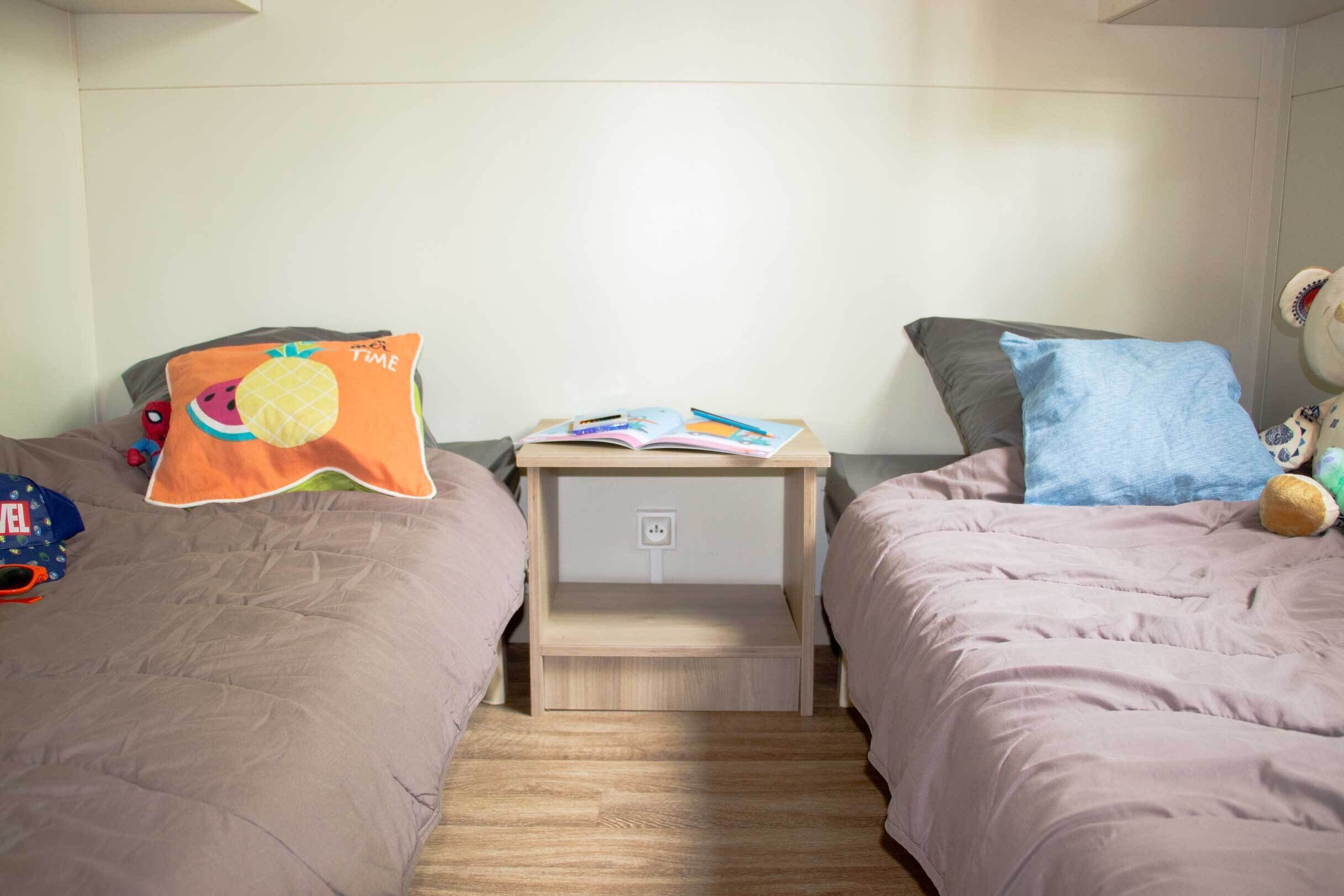 Et soveværelse med to senge, hver med et forskelligt pudedesign, et lille trænatbord med bøger og en lampe, samt en stikkontakt.