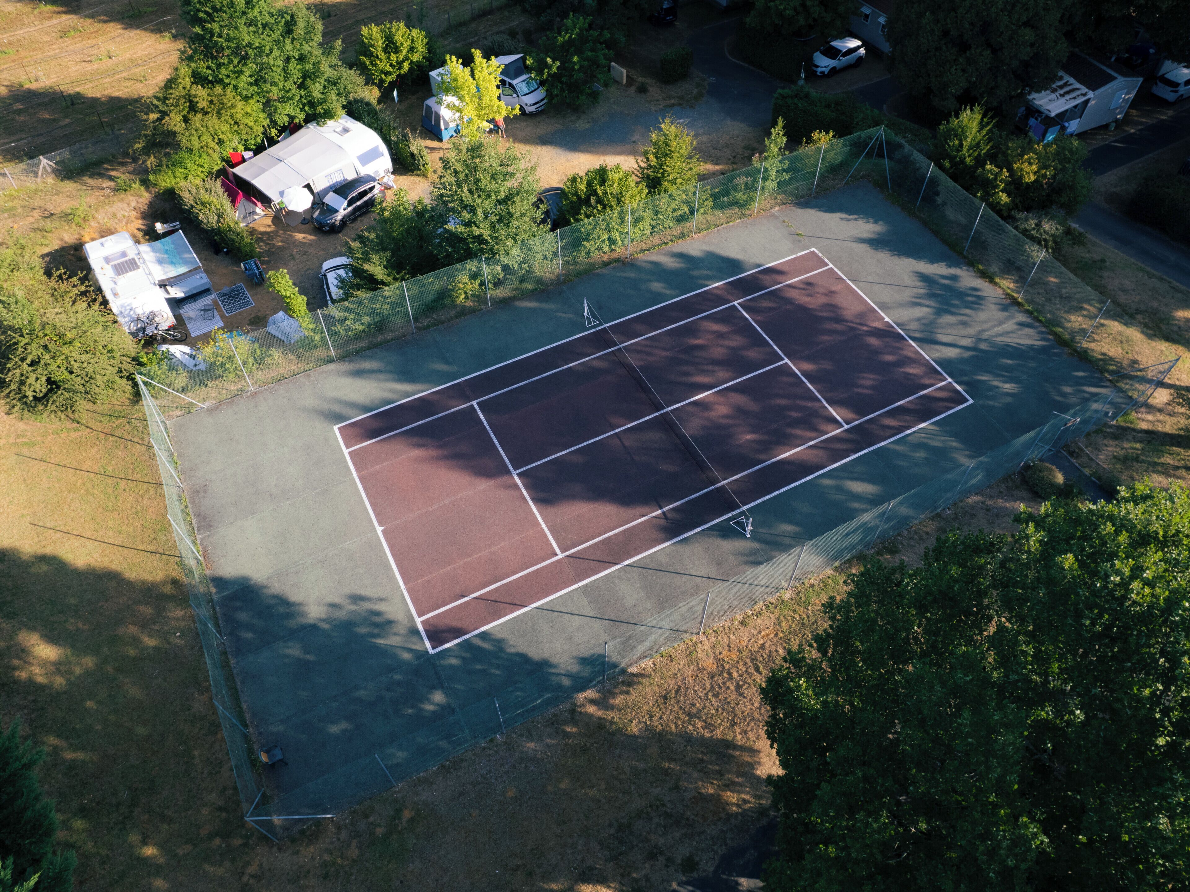Une vue aérienne d’un court de tennis entouré d’arbres et bordé de quelques véhicules garés.