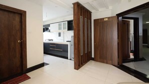 Un couloir d’hôtel moderne, avec des portes en bois, un sol carrelé et un aperçu d’une cuisine équipée de meubles blancs et d’un plan de travail noir.