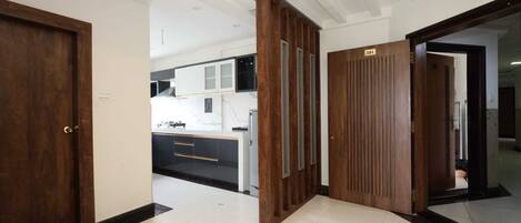 Un couloir d’hôtel moderne, avec des portes en bois, un sol carrelé et un aperçu d’une cuisine équipée de meubles blancs et d’un plan de travail noir.