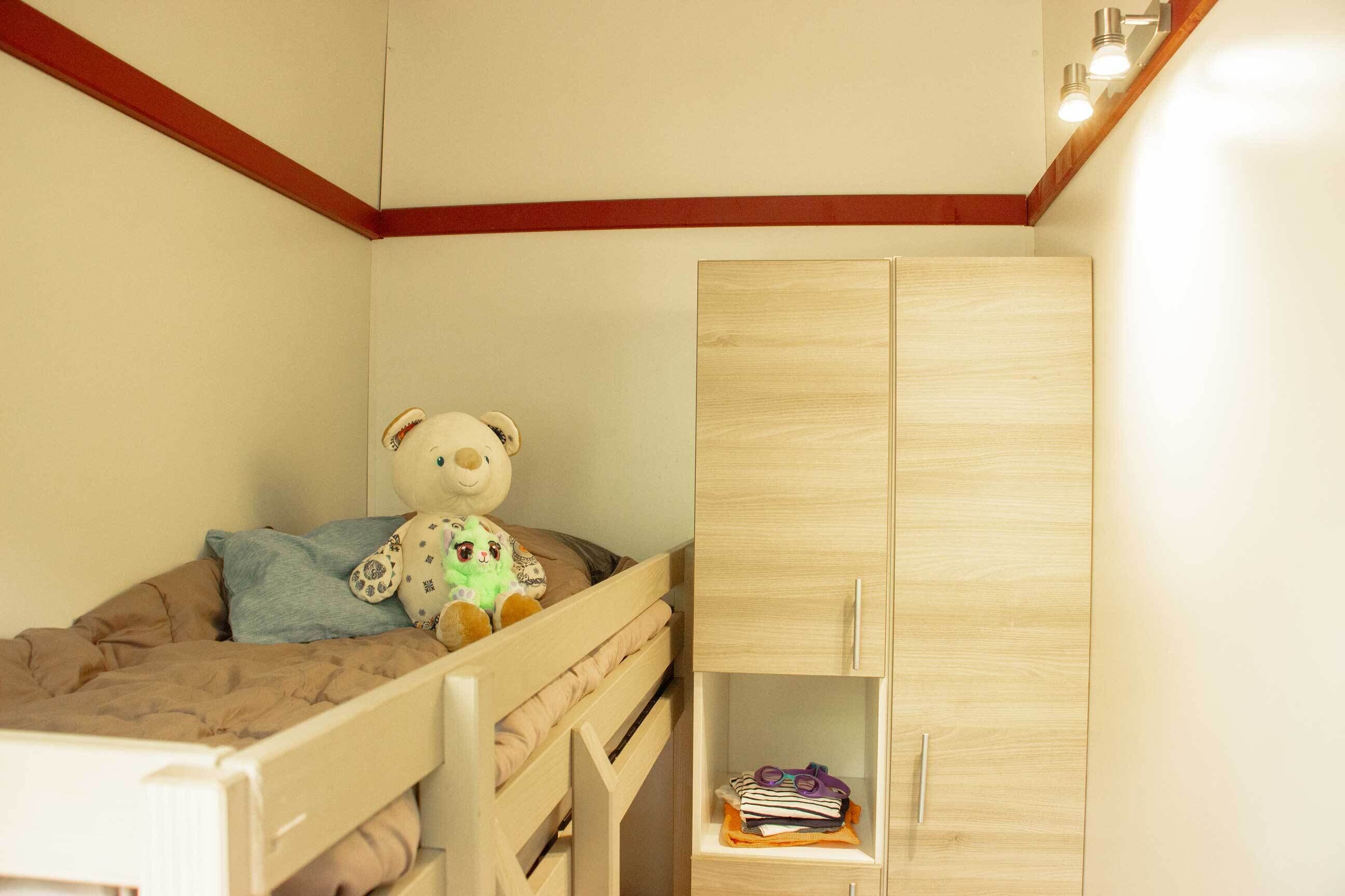 Una cuccetta per bambini con un orsacchiotto, un armadio in legno e uno scaffale con libri e forbici.