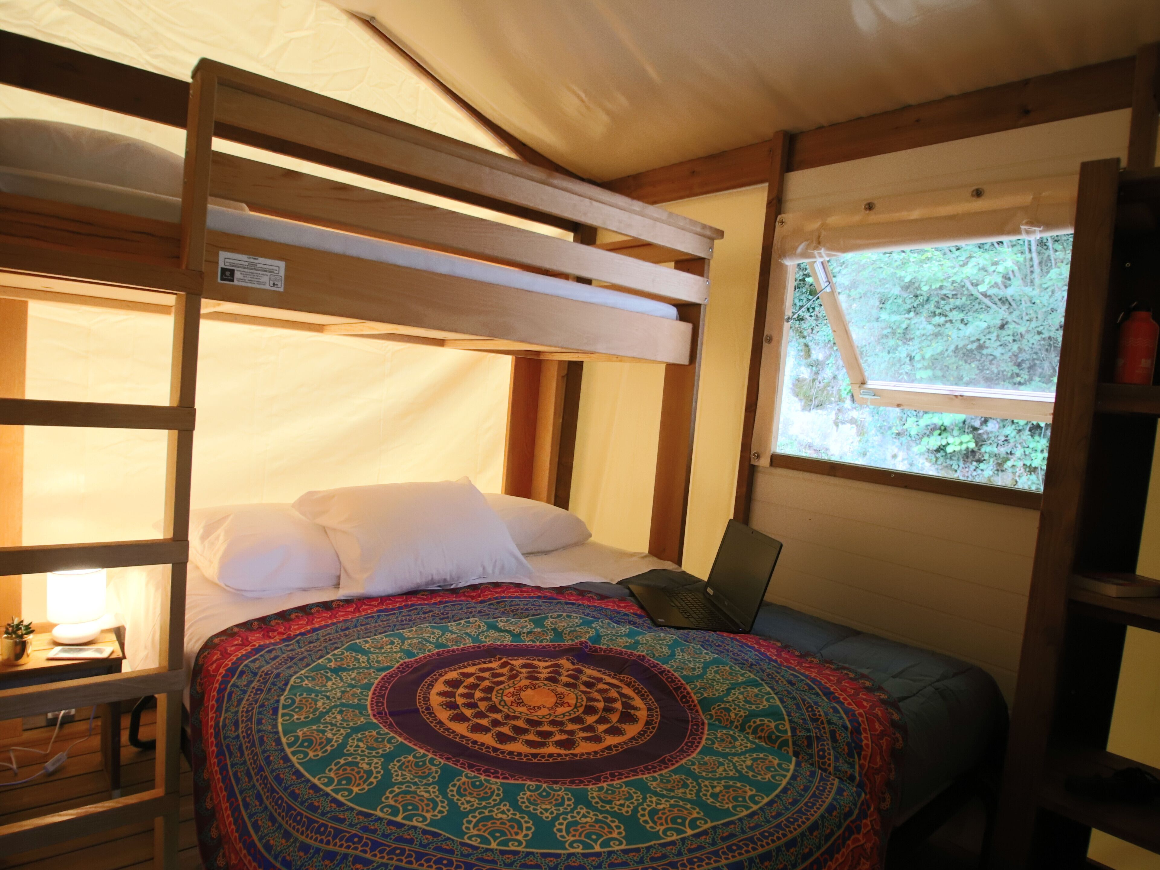 A bunk bed with a colorful tapestry, a bedside lamp, and a window with a view of greenery.