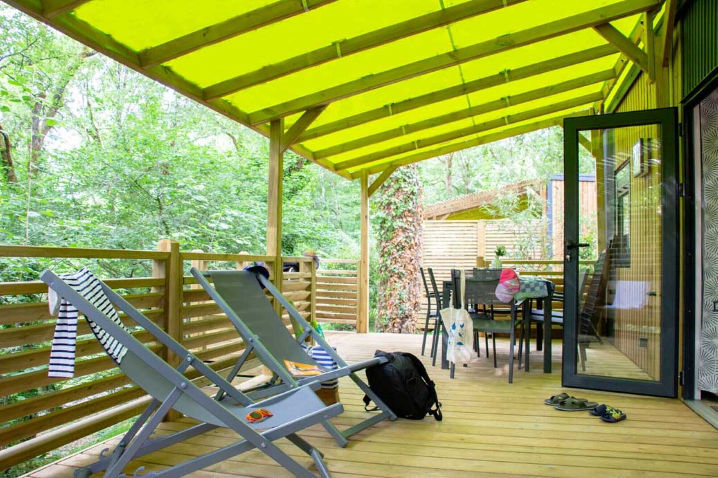 Een overdekt buitengebied met een houten terras, lounge stoelen, een tafel en een glazen deur naar een binnenruimte.