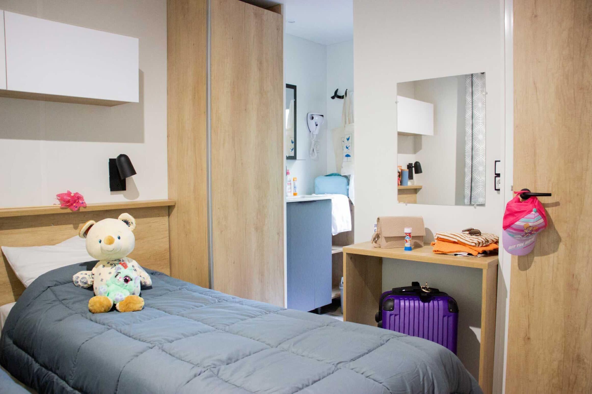 A hotel room with a bed, a bedside table with a suitcase, a teddy bear, a mirror, and a wardrobe.