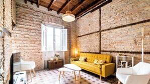 Une pièce avec des murs en briques apparentes, un canapé jaune, des chaises blanches, une petite table et un téléviseur.