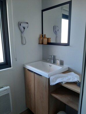 A bathroom with a white sink, wooden cabinet, and a mirror. There are toiletries on a shelf and a hairdryer on the wall. - Truffe - Privilège 3 - 6p (1) (Saint-Geniès)