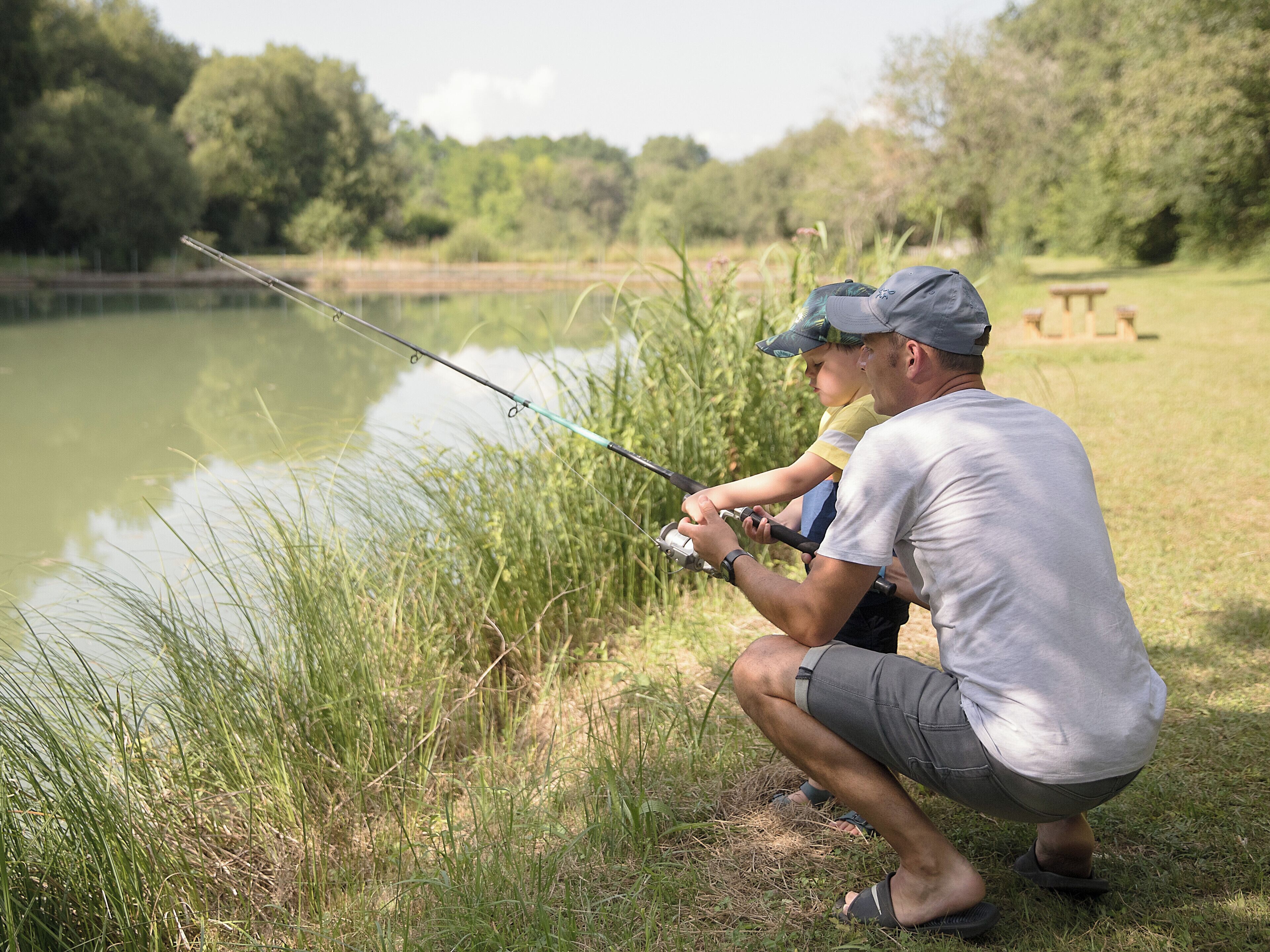 A man and a child fishing by a calm lake surrounded by grass and trees.