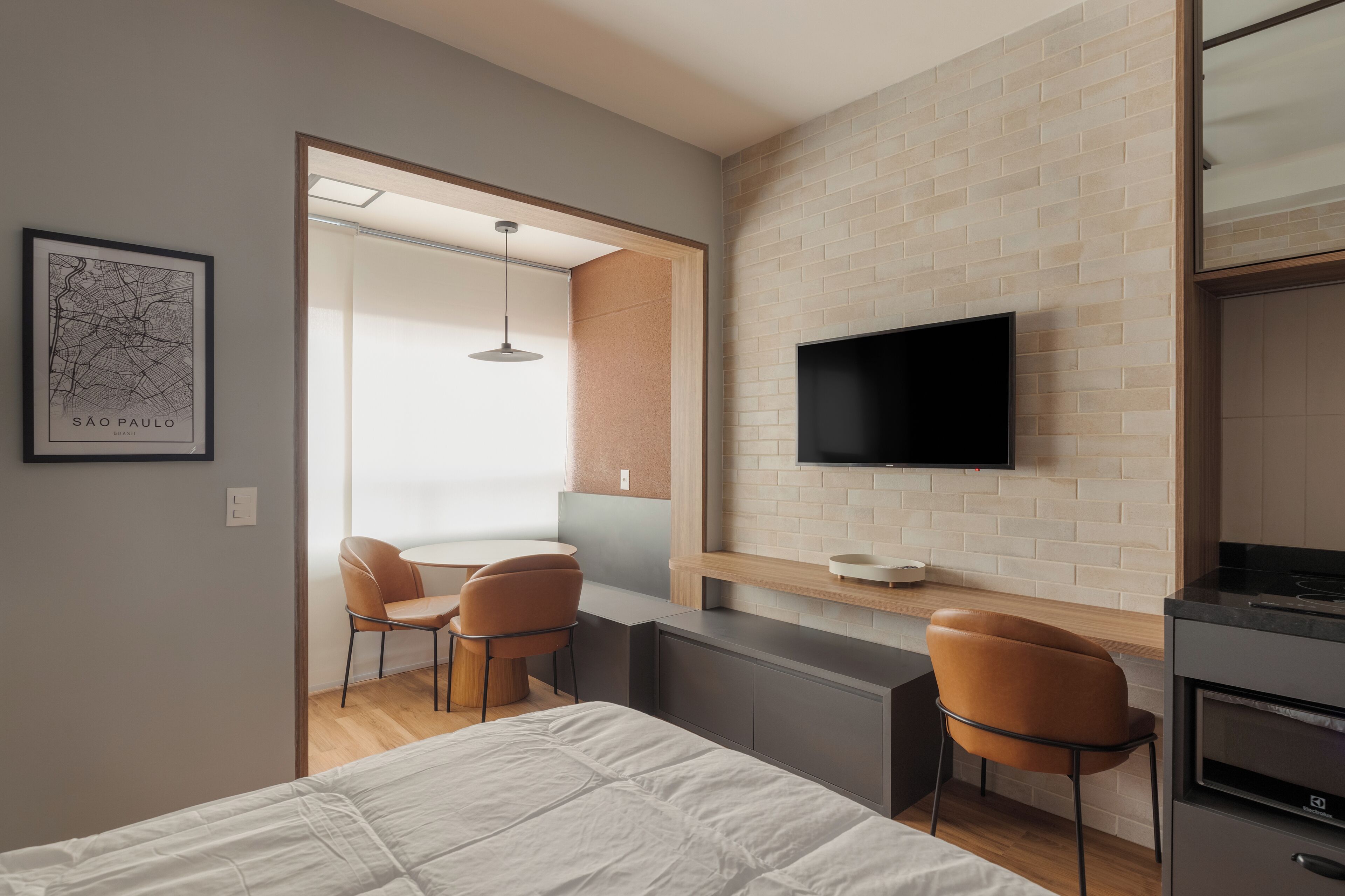 Een moderne hotelkamer met een bed, een televisie bevestigd aan een bakstenen muur, een kitchenette en een zithoek met stoelen.
