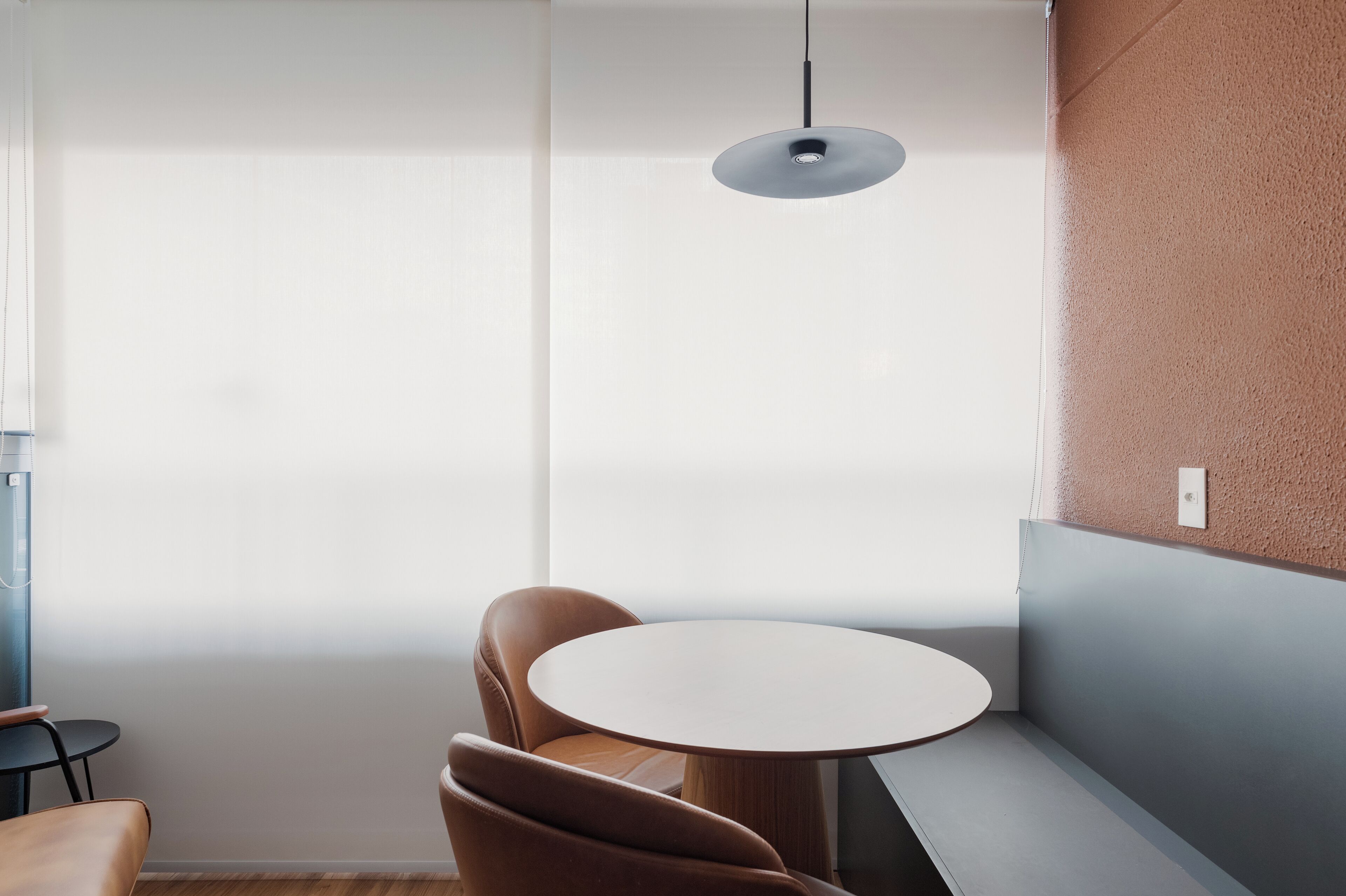 Een moderne kamer met een ronde tafel, twee bruine stoelen, een hanglamp en een groot raam met dunne gordijnen.