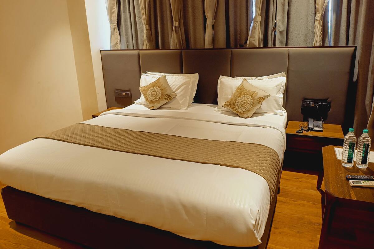 A hotel room with a large bed, two pillows, a bedside table with a phone and water bottles, and a window with curtains.